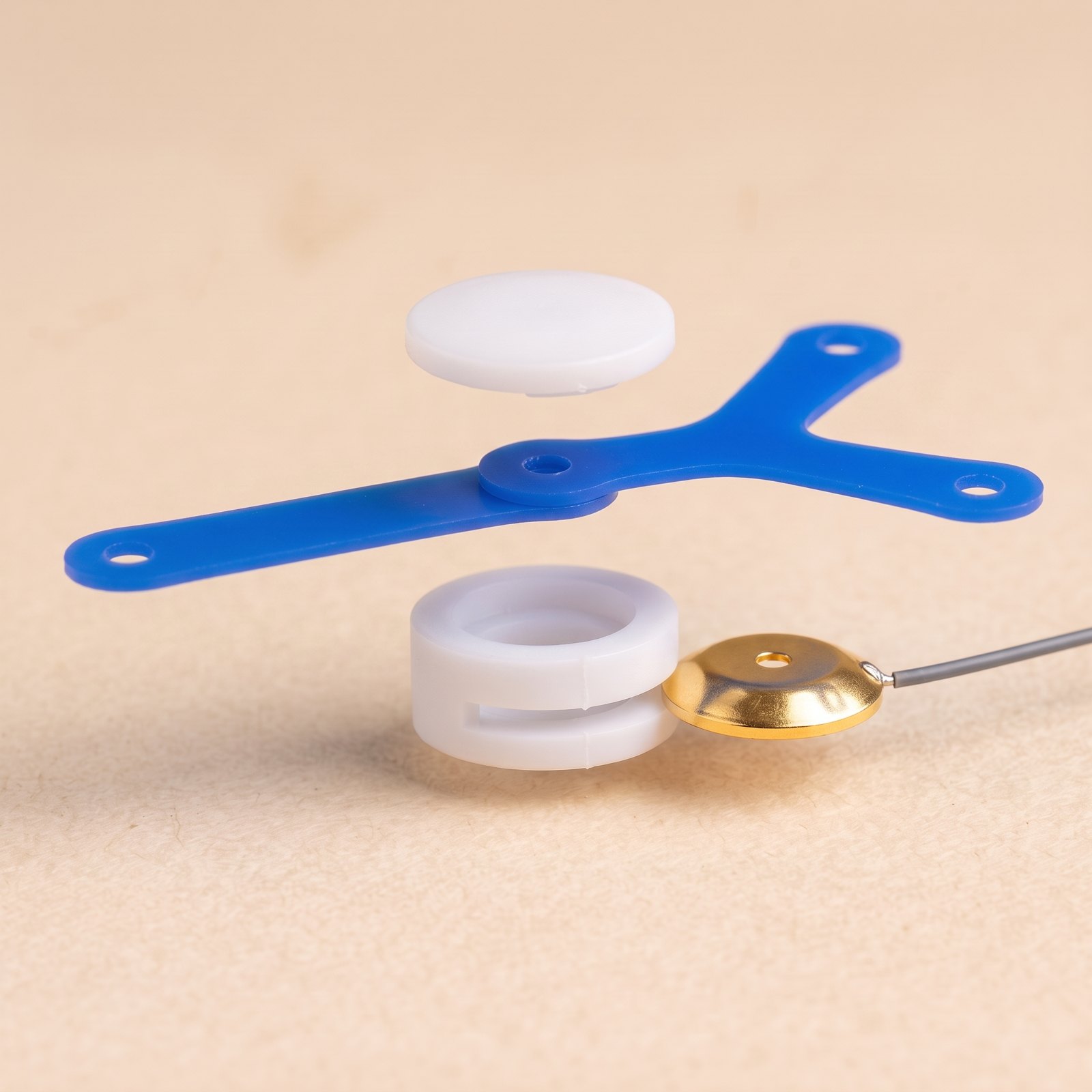 Exploded view: white cap, two thin overlapping silicone strips, a white ring base with a side slot, and a gold cup electrode with its lead lined up to slide into the slot