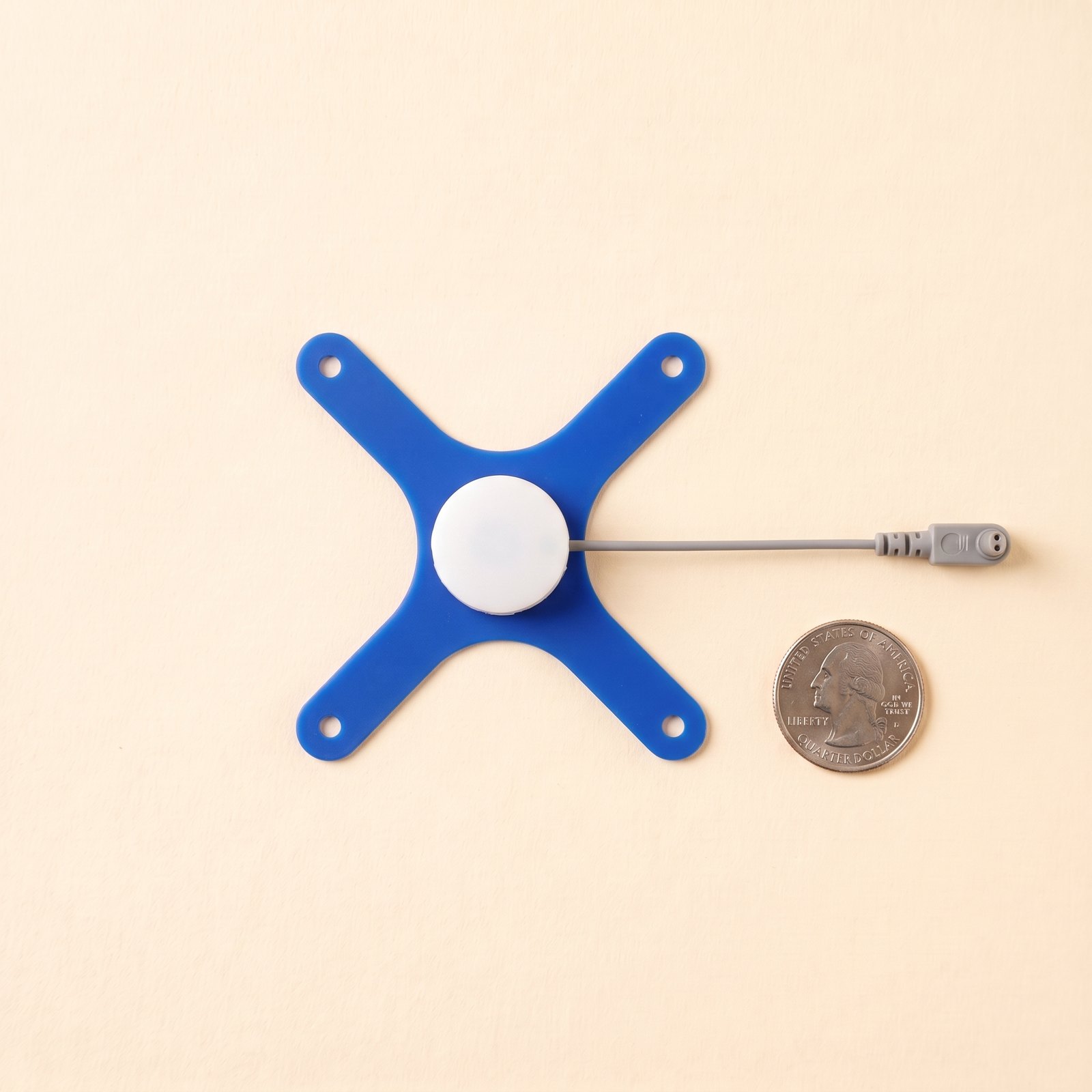 Thin blue X piece with a flat white snap holder at its centre, the lead exiting the holder's slot to a plug, quarter for scale